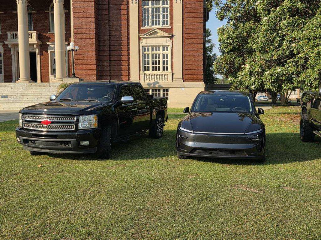 A black pickup truck and a sleek black sedan are parked side by side on grass in front of a large brick building.