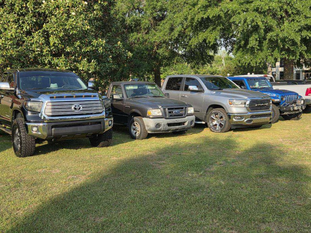 Several pickup trucks and a Jeep are parked on grass under trees, with sunlight casting shadows on the scene.