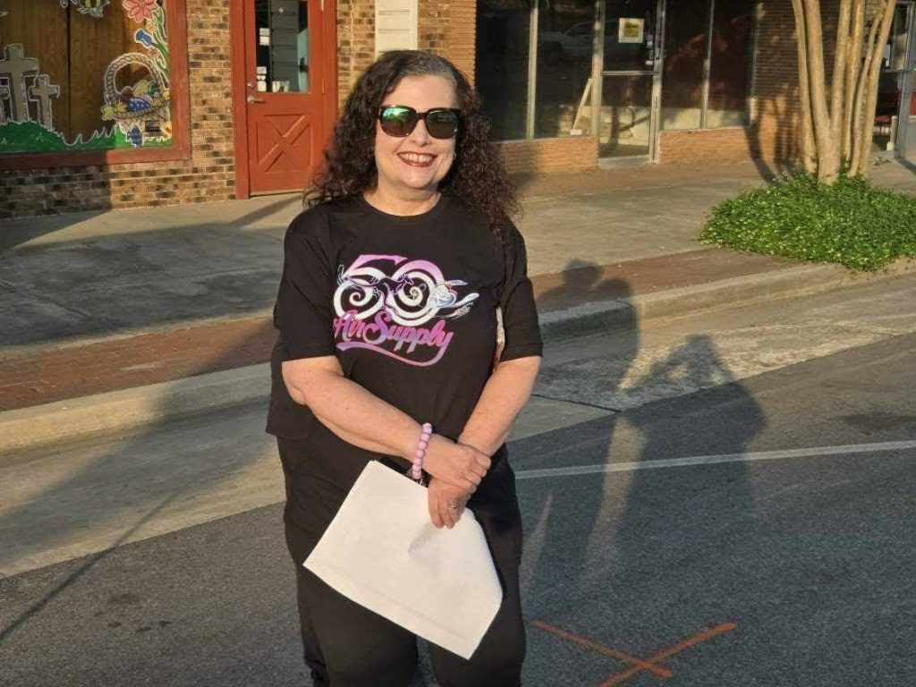 A woman wearing sunglasses and a "50 Air Supply" t-shirt stands on a street holding a piece of paper, with buildings and painted windows behind her.