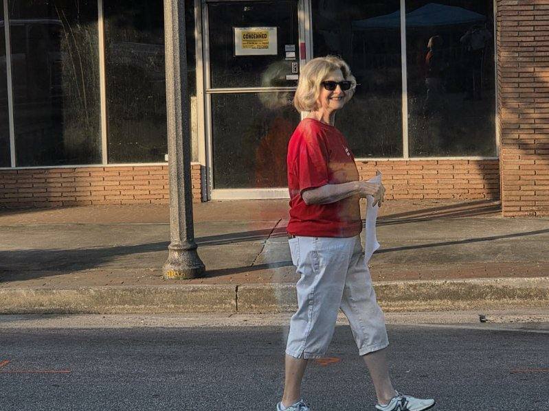 older woman in a red shirt and white pants walks on a city sidewalk, smiling, holding paper, with a "condemned" sign visible in the building behind her.