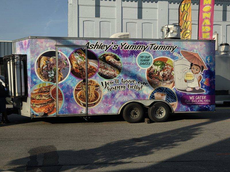Colorful food truck labeled "Ashley's Yummy Tummy" with images of food, cartoon bear, and catering contact information, advertising sauces and the slogan, "You'll have one happy belly!"