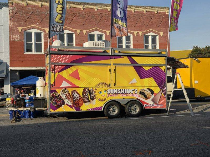 A brightly colored food trailer labeled "Sunshine's Sweet Creations" with dessert images is parked on a street in front of a brick building.
