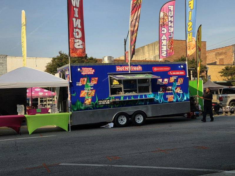 A blue food truck with colorful banners reading "Wings," "Fish," "Tenders," and "Steak," parked at an outdoor event with nearby tents and tables.