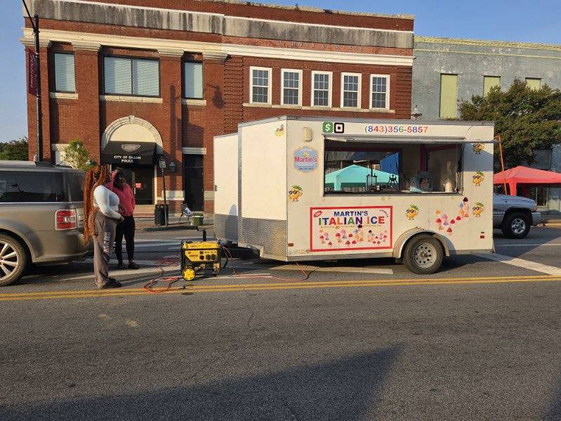 A white food truck labeled "Martin's Italian Ice" is parked on a street with two people standing nearby and a generator on the ground.