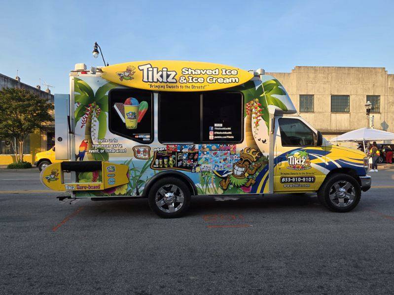 A colorful Tikiz shaved ice and ice cream truck parked on the street, decorated with tropical designs and menu images on the side.