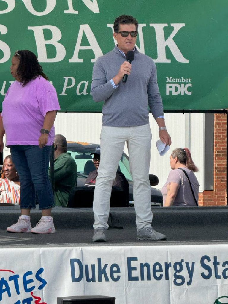 A man wearing sunglasses speaks into a microphone on an outdoor stage, with people and a green event banner in the background.