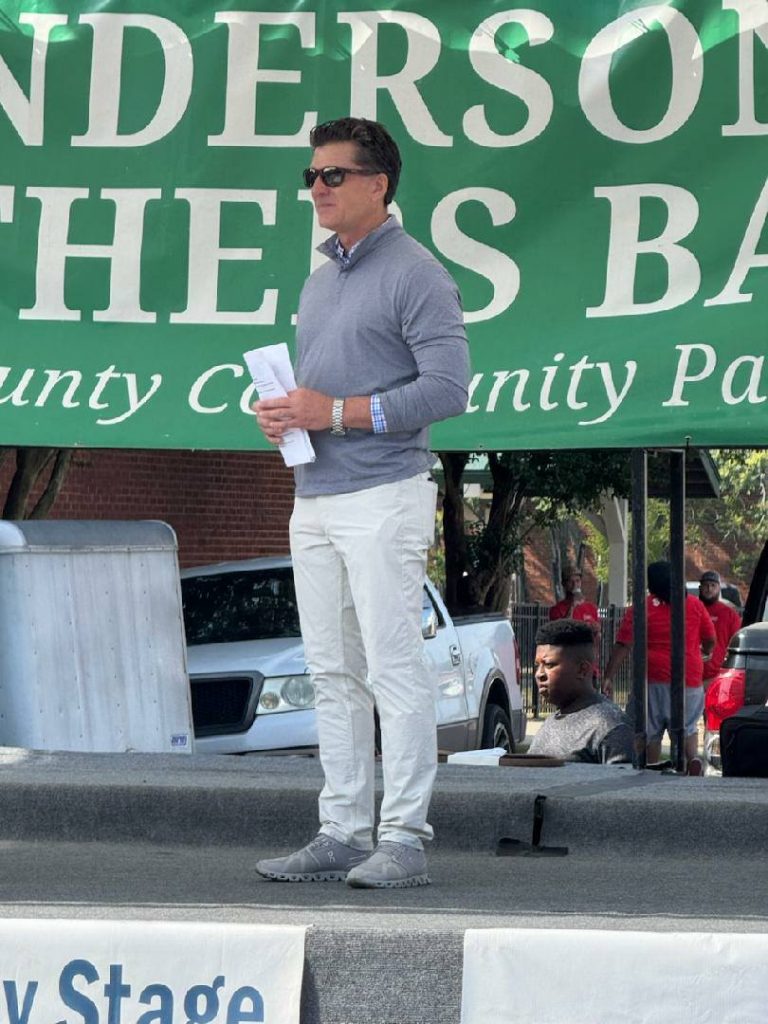 A man in sunglasses stands on an outdoor stage, holding papers, with a green event banner and parked vehicles in the background.