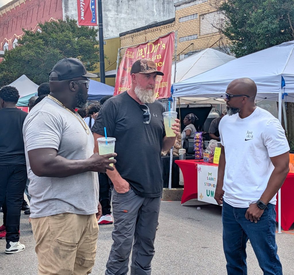 Three men stand outdoors at a street festival, holding drinks and talking. Vendor booths and people are visible in the background under tents.