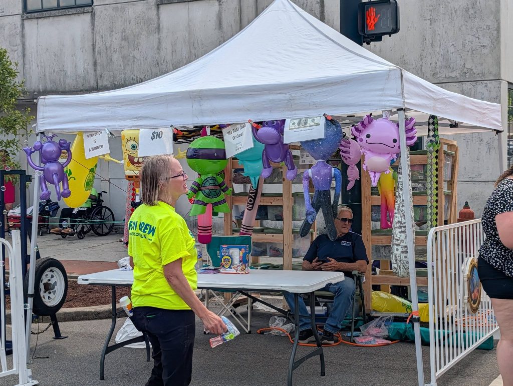 Outdoor carnival booth with inflatable toys, signs, and two people under a white tent; one person standing, another sitting behind a table. "Everybody is a winner" sign visible.