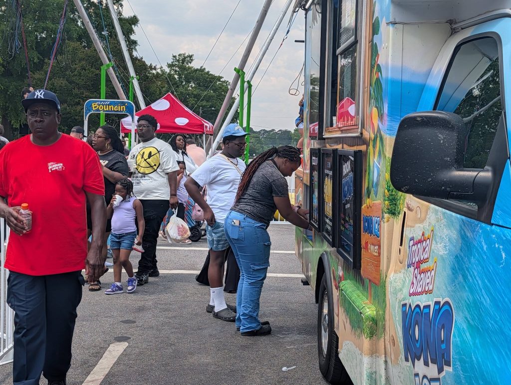 People standing near a colorful shaved ice truck at an outdoor event, with children and adults walking and waiting in line, and tents and rides in the background.