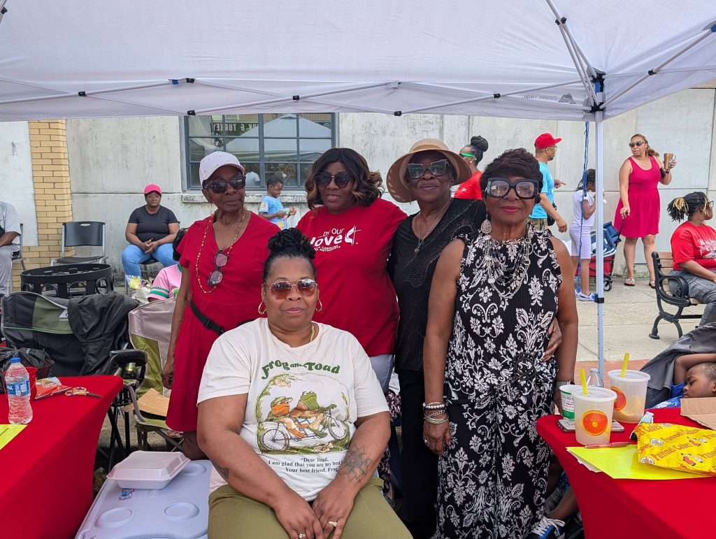 Five women posing under a canopy at an outdoor event, with tables, drinks, and people in the background.