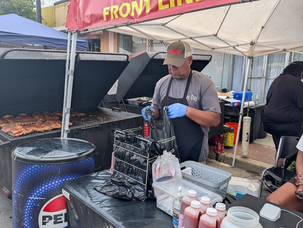 A man in a cap and gloves prepares food at a barbecue stand, with ribs grilling on a large smoker under a canopy at an outdoor event.