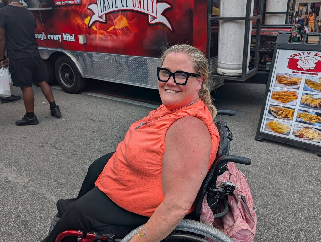 A smiling woman in a wheelchair wearing glasses and an orange shirt at a food truck festival, with menu displays and people in the background.