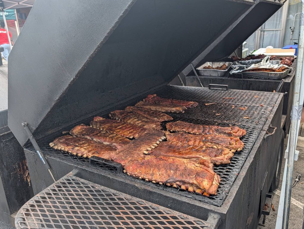 Several racks of seasoned pork ribs cooking on a large outdoor barbecue smoker with the lid open.
