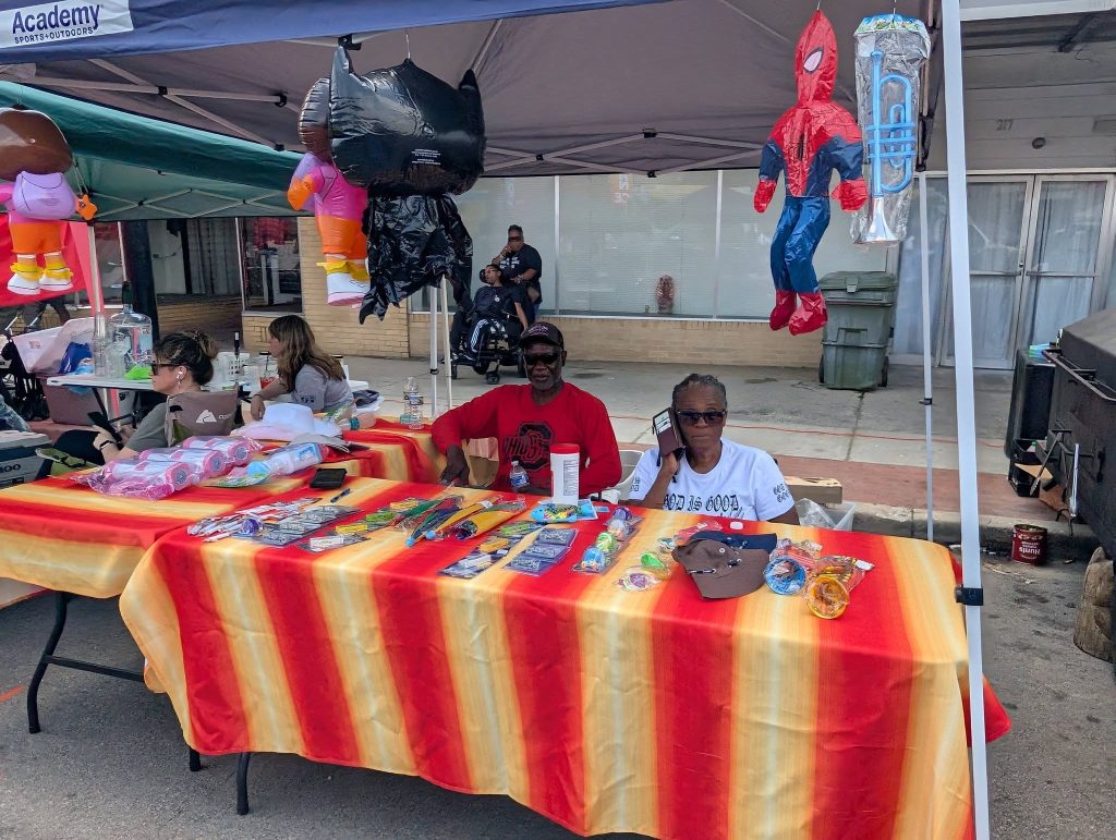 People sit under a canopy at a street fair booth with toys and colorful items on red-yellow striped tables, inflatable characters hanging overhead.