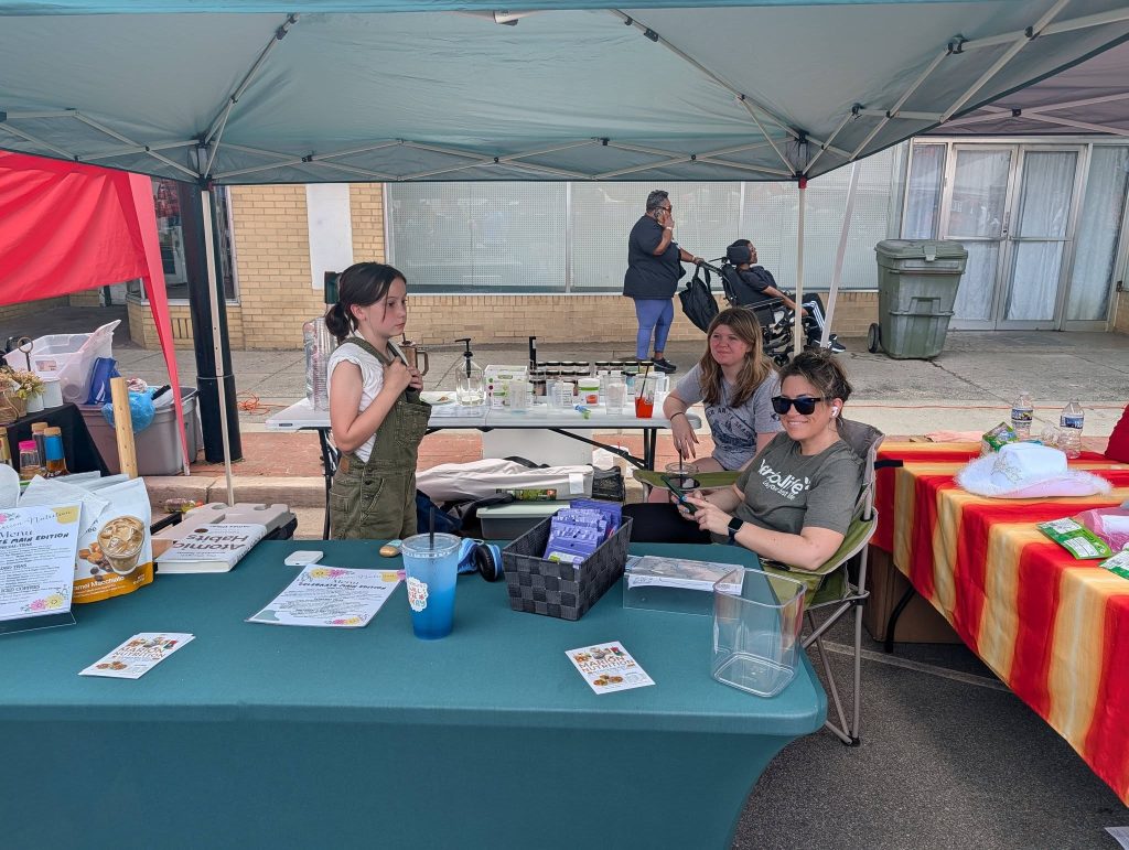 Three women at a market booth under a canopy, with informational flyers, products, and drinks on display, while people walk and sit in the background on the sidewalk.