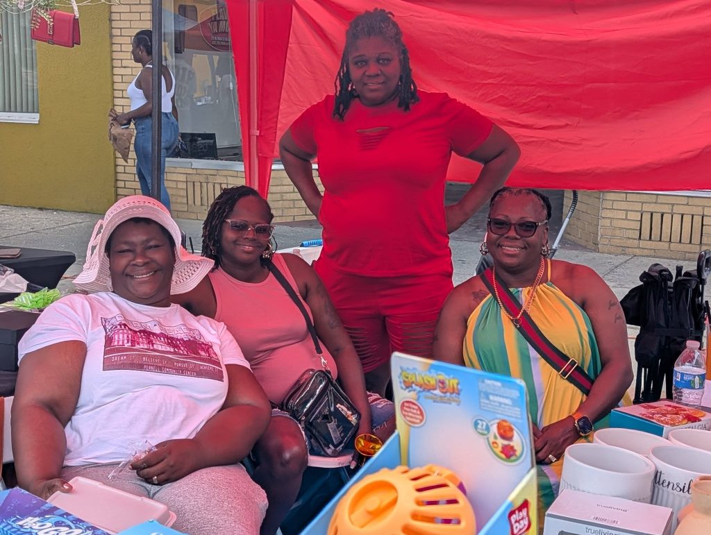 Four women sitting and smiling under a red canopy at an outdoor event, surrounded by mugs, toys, and other items on a table.
