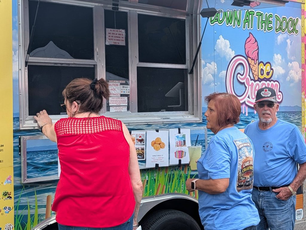 Three people stand at an ice cream truck, with one woman in red ordering while two others in blue shirts wait by the menu.