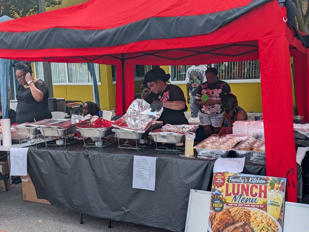 People serving food under a red canopy at a street food stall, with trays of dishes and a lunch menu sign displayed in front.