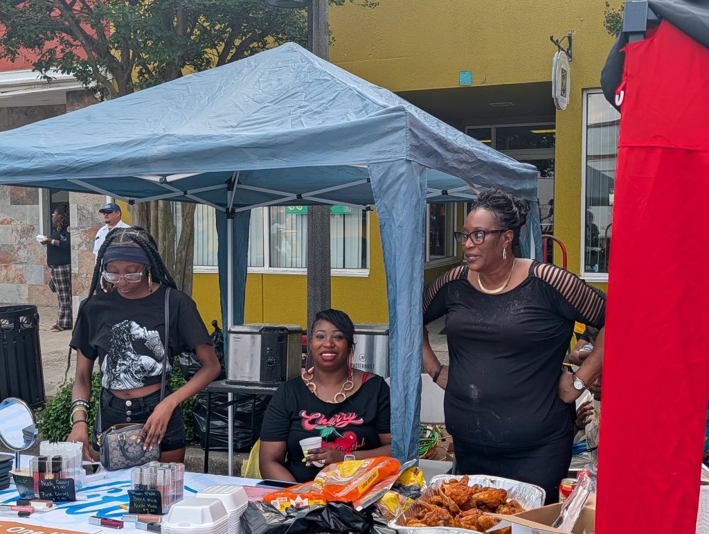 Three women at a street food stall under a canopy, with trays of food, cosmetics, and assorted items displayed on the table.