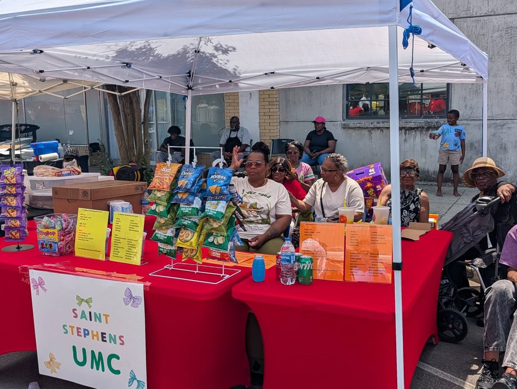 A group of people sitting behind a red table with snacks for sale under a white canopy at a Saint Stephens UMC outdoor event.