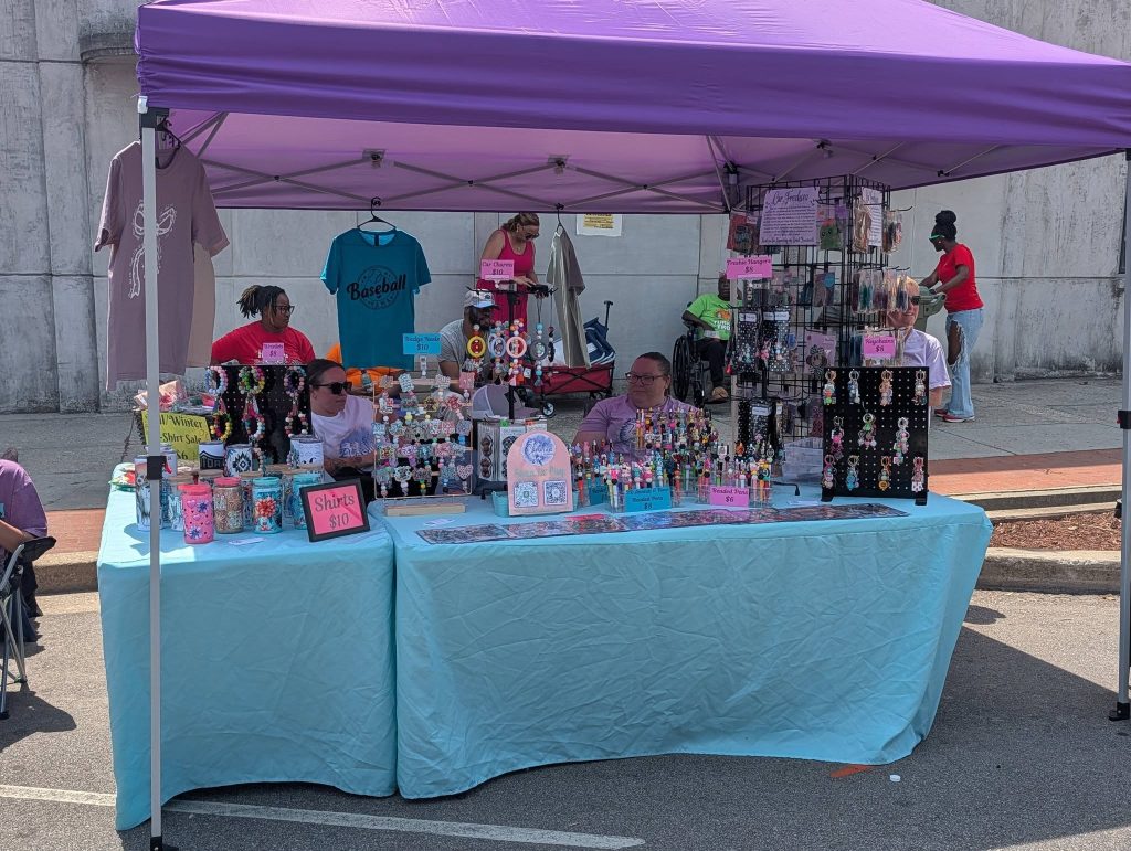 Outdoor market booth with two people selling handmade jewelry, shirts, and crafts under a purple canopy. Tables display colorful items and price signs; passersby in the background.