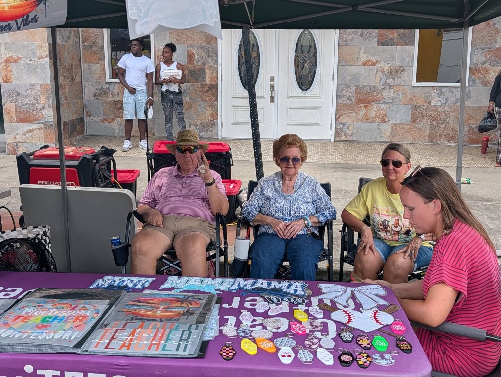 Four people sit at a booth with various crafts and signs on display, while two others stand in the background on a sidewalk by double doors.