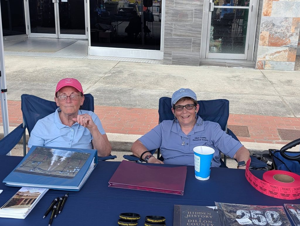 Two women sit at a table outside with historical society materials, raffle tickets, brochures, and books displayed in front of them.