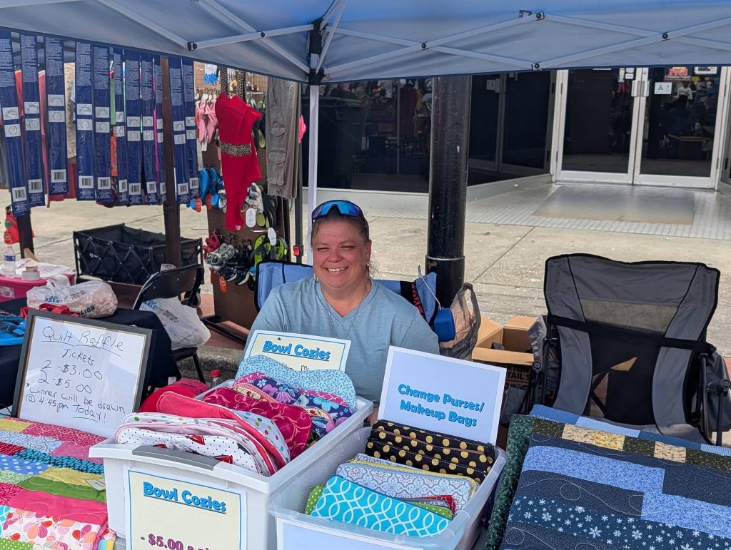 Smiling woman sits at a craft fair booth selling bowl cozies, quilts, and makeup bags under a blue canopy. Signs and colorful fabric items are displayed.