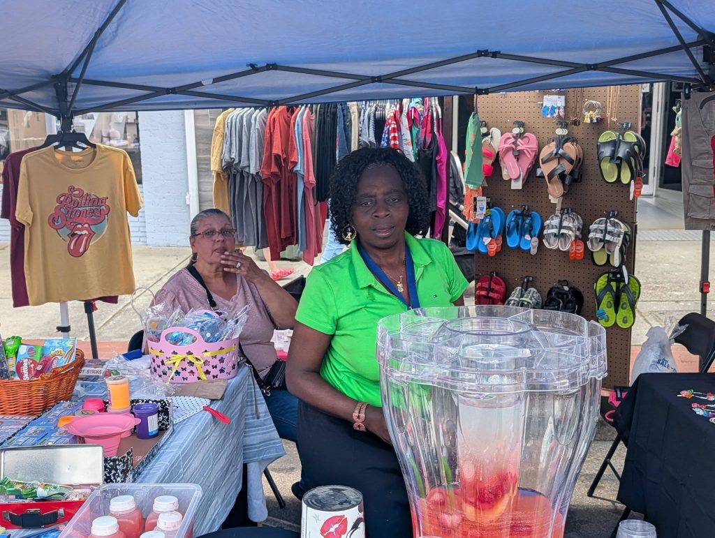 Two women sit at a market stall selling drinks, snacks, shirts, and sandals under a blue canopy on a city sidewalk.