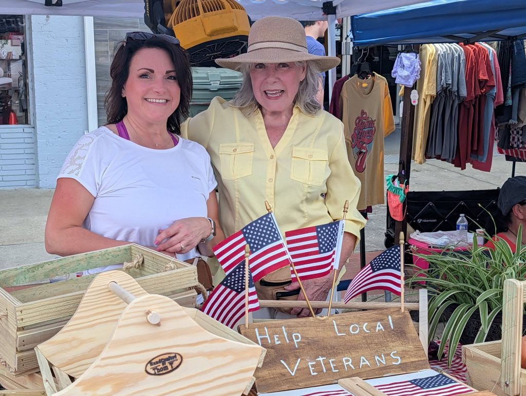 Two women stand behind a market booth with American flags and a sign reading "Help Local Veterans," surrounded by handmade wooden crafts and crates.
