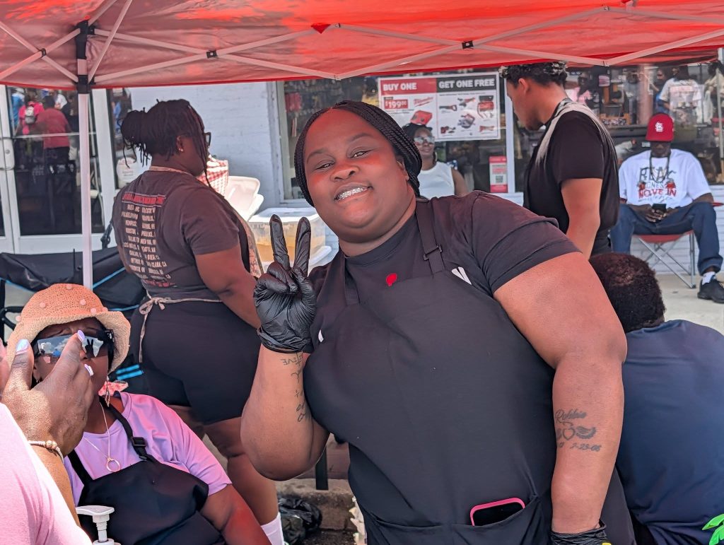 Woman in black apron smiles and gives peace sign under red canopy; people around her are seated or standing at an outdoor event.