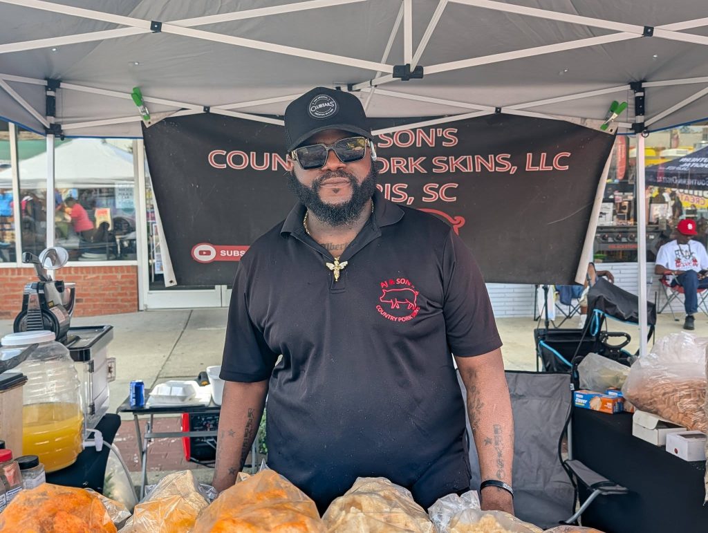 Man wearing sunglasses and a black shirt stands behind a market stall with bags of pork skins under a tent, outdoor setting.
