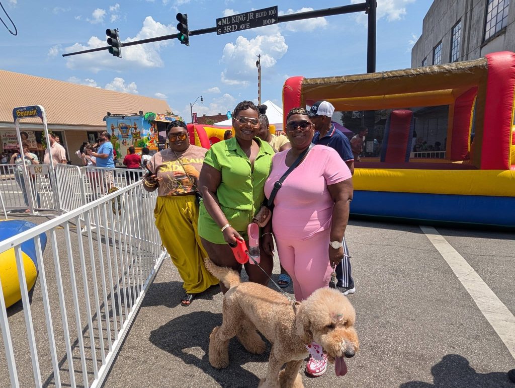 Three women posing with a large poodle at a street fair, with bounce houses and carnival booths in the background on a sunny day.