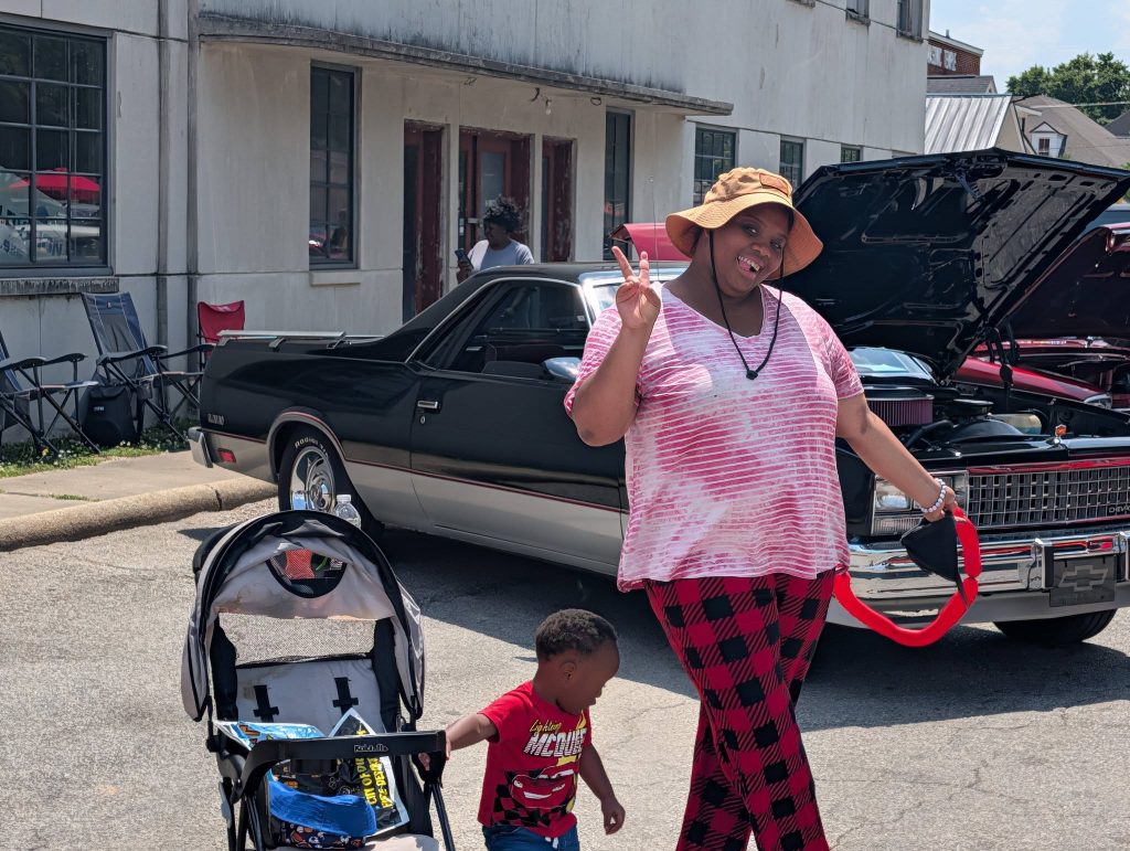 Woman posing with peace sign near classic car, wearing a hat and pink clothes, holding a leash, with a child and stroller nearby at an outdoor event.