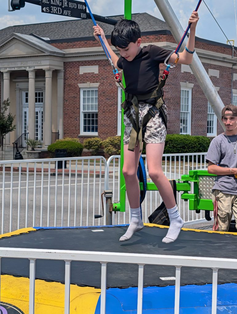 A boy in a harness bounces on a trampoline at an outdoor event, with a brick building and street signs visible in the background.