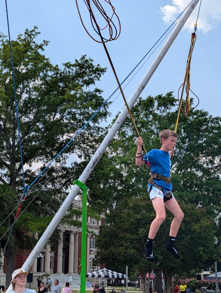 Boy in harness bounces on outdoor bungee trampoline at a fair, surrounded by trees and people, with a building visible in the background.