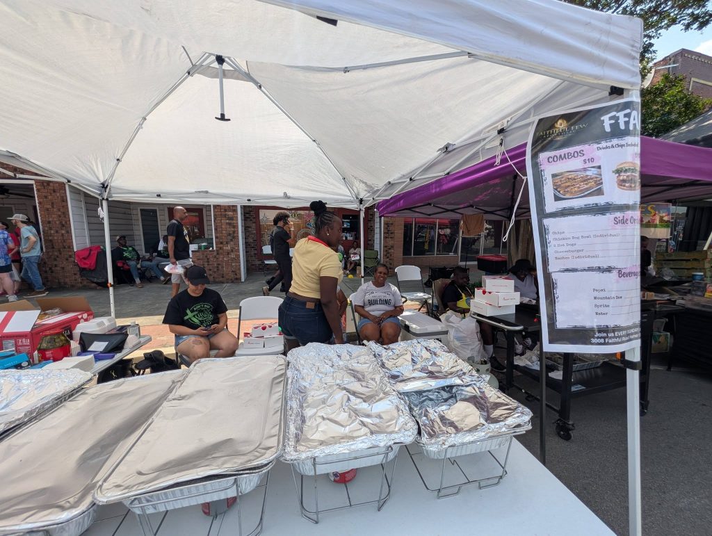 People sitting under a white canopy at a food stand with covered trays, a combo menu sign, and items on tables at an outdoor market or event.