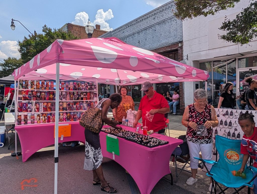 People browsing and selling items under a pink polka dot tent at an outdoor street market, with colorful cups and jewelry displayed on tables.