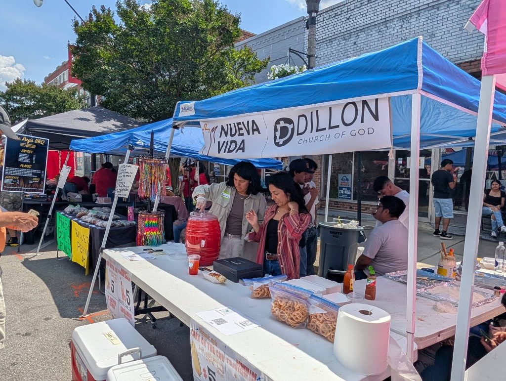 People at a street fair stand under a blue tent labeled "Nueva Vida Dillon Church of God," selling drinks and snacks, with other vendor booths nearby.