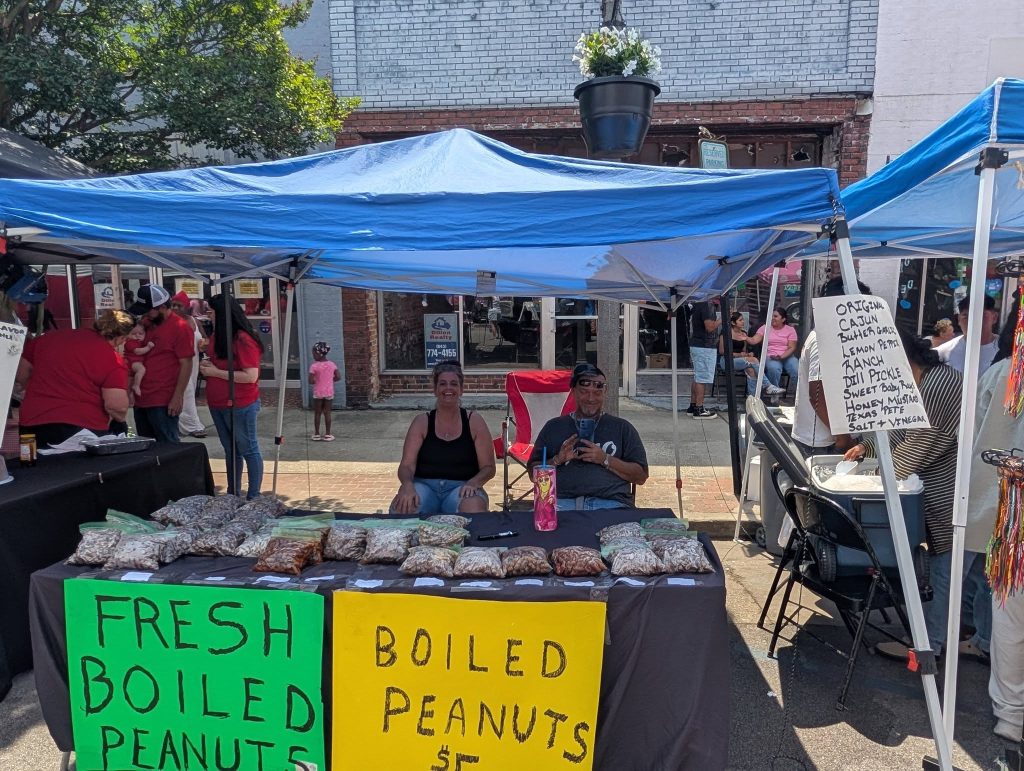 Two people sit under a blue tent at a street market, selling bags of boiled peanuts, with bright signs and a menu of flavors displayed.