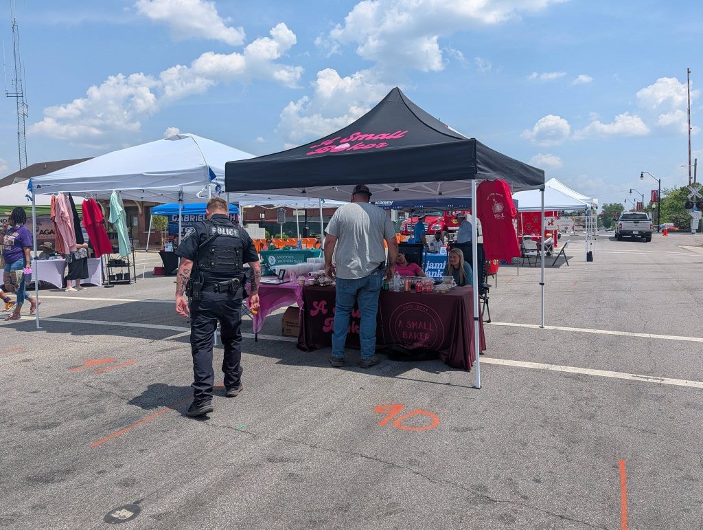 Outdoor market with vendor tents, people browsing booths, clothing displays, police officer walking, and a blue sky with clouds in the background.