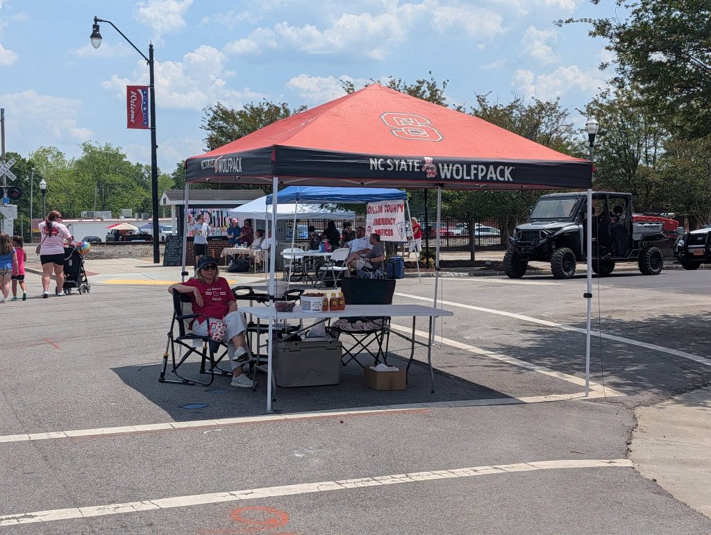 A person sits under a red tent labeled "Wolfpack" in a parking lot with tables and items, other tents and vehicles in the background.