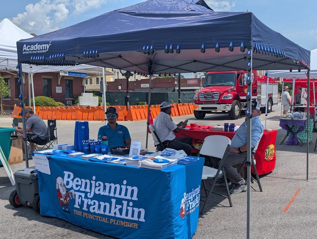 Outdoor event booth with "Benjamin Franklin The Punctual Plumber" table, pens, cups, and pamphlets. People sitting under tents, orange barriers, and fire truck in background.