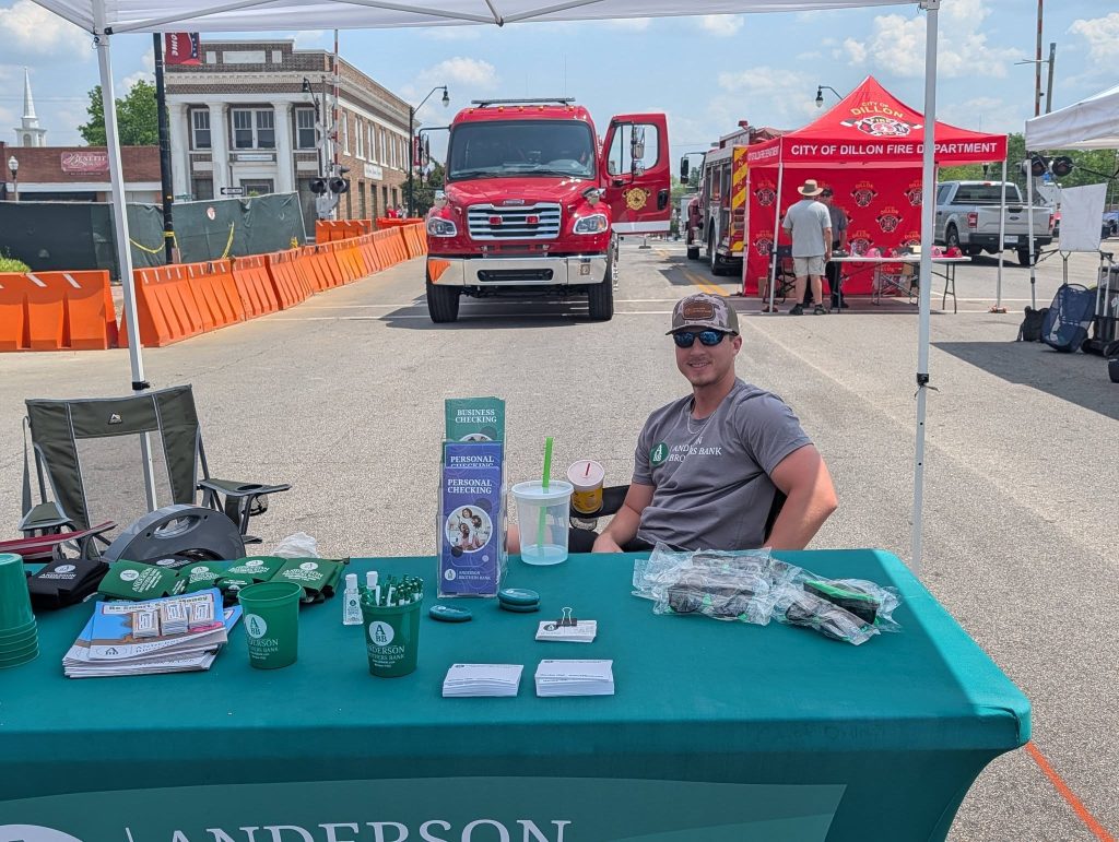 Man sits at a promotional booth for Anderson Brothers Bank at a street event, with fire trucks and a City of Dillon Fire Department tent in the background.