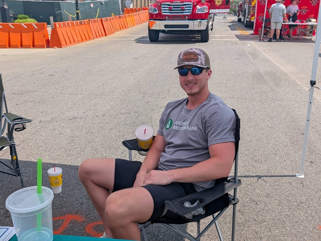 A man in sunglasses and a cap sits in a folding chair at an outdoor event, smiling, with drinks beside him and emergency vehicles in the background.