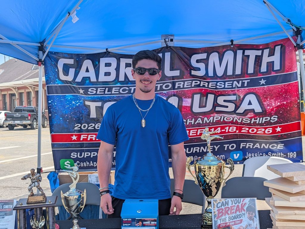 A man in a blue shirt stands behind a table with trophies and wooden boards, promoting Team USA at a martial arts event under a blue canopy.