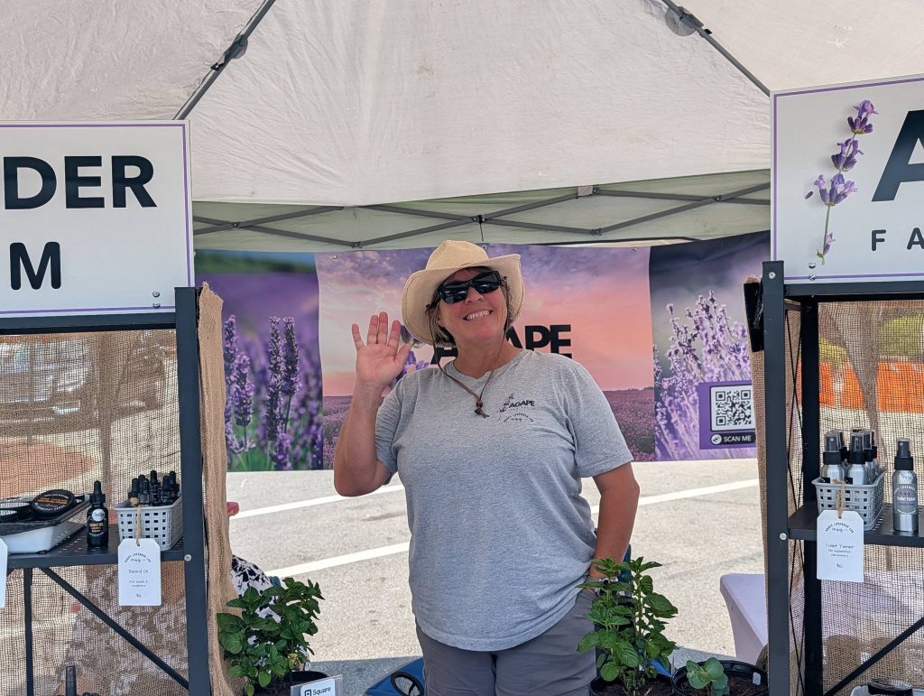 Smiling woman in a hat waves at a lavender farm market stall, with lavender products and plants displayed under a white tent.