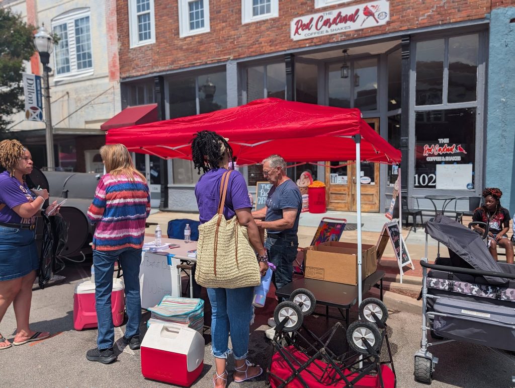 People gather under a red canopy at a street fair in front of The Red Cardinal Coffee & Shakes, with coolers, tables, and a stroller visible.