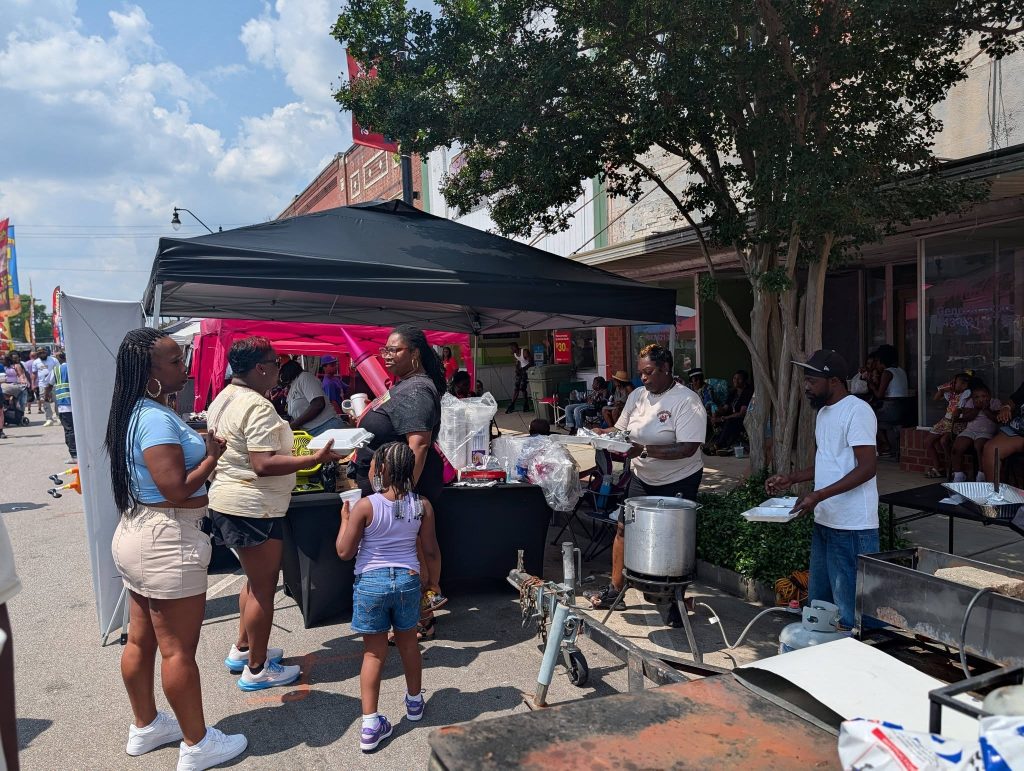 People gather at an outdoor food stand on a sunny day, chatting, cooking, and serving meals under tents on a city street.